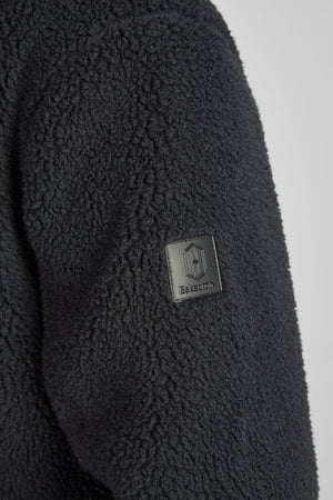 L'image montre un gros plan d'une veste en tissu bouclé noir, probablement en matière polaire ou en laine synthétique, offrant une texture douce et moelleuse. Sur le côté gauche de la veste, près du bas, il y a un patch en cuir noir cousu. Ce patch affiche un logo distinctif composé d'éléments géométriques, notamment un carré avec deux lignes verticales à l'intérieur, et une petite partie de texte en dessous qui semble indiquer une marque ou un logo. Le tissu de la veste présente une texture...