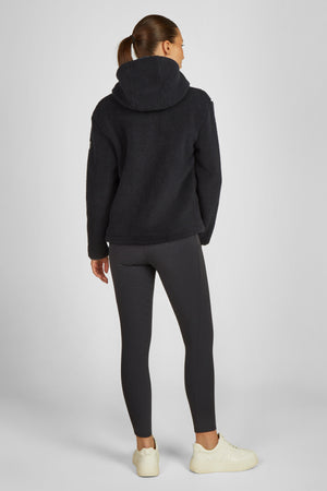 L'image représente une femme vue de dos, se tenant debout. Elle porte une tenue décontractée et confortable, adaptée pour le sport ou les activités de loisirs. Voici une description détaillée de son apparence et de sa tenue : 1. **Vêtements**: - **Haut**: La femme porte un sweat-shirt noir à capuche. La texture semble moelleuse et douce, probablement en tissu polaire, offrant une sensation de confort et de chaleur. Le sweat-shirt a une coupe décontractée et est doté d'une capuche bien...