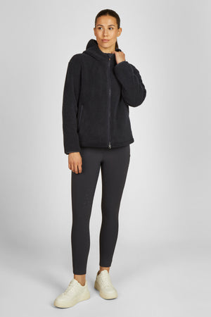 Dans l'image, une femme se tient debout contre un fond blanc uni, ce qui met en valeur sa tenue sportive élégante. Elle porte une veste en tissu moelleux et pelucheux de couleur anthracite qui semble douce et confortable, parfaite pour les journées fraîches. La veste est zippée jusqu'à son cou et dispose d'une capuche, lui offrant à la fois style et fonctionnalité. Sous sa veste, elle porte un haut ajusté et des leggings de la même couleur anthracite. Les leggings sont de type taille haute,...