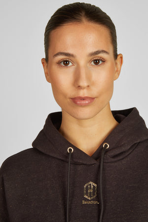 Cette image montre une femme photographiée en position frontale. Son expression faciale est neutre. Elle a les cheveux tirés en arrière de manière soignée, dégageant son visage et révélant ses traits. Elle porte un hoodie noir avec une capuche doublée de noir également. Le hoodie semble être de marque, car il arbore un petit logo brodé doré sur le côté gauche de la poitrine. Le logo est composé de deux lettres "H" superposées, avec le mot "Eskadron" écrit en dessous. L'arrière-plan de la...