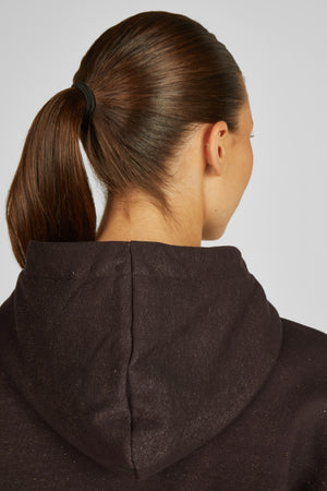L'image montre une personne vue de dos, se concentrant principalement sur la partie supérieure de son corps. La personne a des cheveux bruns foncés attachés en une queue de cheval haute avec un élastique noir. Le visage est partiellement visible de profil. Elle porte un vêtement noir, probablement un sweat-shirt à capuche, qui est légèrement poussiéreux ou parsemé de petites taches blanches. Le tissu du sweat-shirt semble être de texture douce et plutôt épais. Le fond est blanc et neutre,...