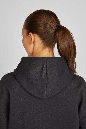 L'image montre l'arrière d'une personne portant un sweat-shirt noir à capuche. La capuche du sweat-shirt est bien visible et repose sur les épaules. La personne a les cheveux attachés en queue de cheval haute, maintenue par un élastique noir. Les cheveux sont bruns et ont l'air lisses et bien coiffés. Le sweat-shirt semble être fait d'un tissu doux et confortable, probablement du coton ou un mélange similaire. L'éclairage est uniforme, mettant en valeur les détails de la tenue et de la...