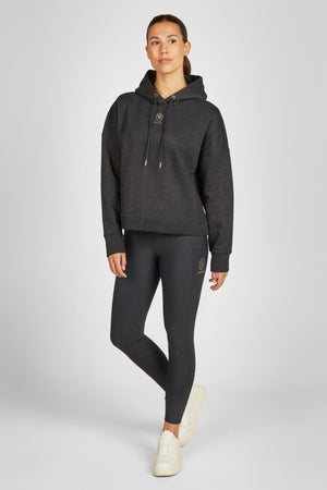 Sur cette image, on peut voir une femme debout dans une pose décontractée. Elle est vêtue d'une tenue de sport élégante et confortable. Elle porte un sweat à capuche noir avec une texture légèrement brillante, ce qui donne un effet subtilement scintillant à l'ensemble. Le sweat a des manches longues et une capuche avec des cordons de serrage. Sur la poitrine à gauche, il y a un petit logo doré, qui semble être la marque de la tenue. Le sweat est ample et confortable, idéal pour des...