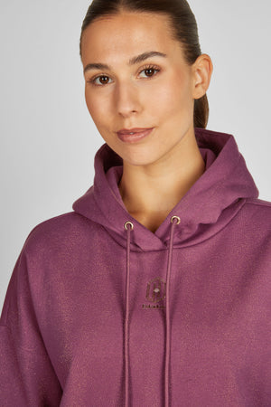 L'image représente une femme portant un sweat-shirt à capuche de couleur violette. Elle a les cheveux tirés en arrière, dégageant son visage. Le sweat-shirt possède une capuche avec des cordons de serrage de couleur similaire, qui sont légèrement visibles sur ses épaules. Sur le côté gauche de la poitrine du sweat-shirt, il y a un petit logo en forme de bouclier avec des détails dorés et une inscription. Le fond est blanc uni, ce qui met l'accent sur la personne et le vêtement qu'elle porte....