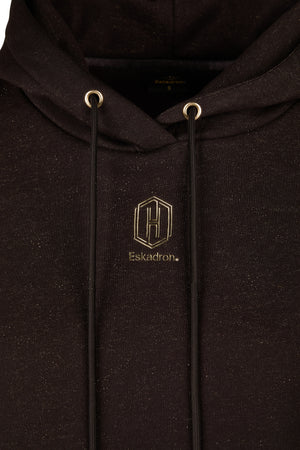 L'image montre un sweat-shirt noir avec une capuche. Le tissu du sweat-shirt semble contenir de légères particules dorées scintillantes, lui donnant un aspect subtilement brillant. Le sweat-shirt présente un logo brodé dans une couleur dorée, situé au niveau du cœur. Le logo consiste en un hexagone contenant la lettre "H" en son centre, avec le mot "Eskadron" écrit en dessous. Les cordons de la capuche sont noirs et passent à travers des œillets dorés, renforçant ainsi l'esthétique générale...