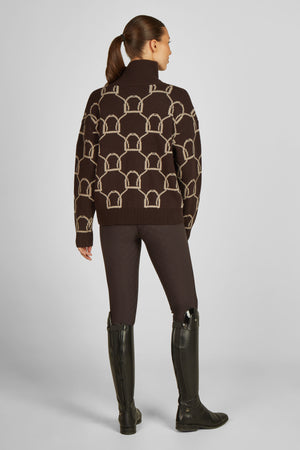 L'image montre une femme vue de dos, portant une tenue élégante et décontractée. Elle porte un pull à col roulé marron avec un motif géométrique distinct en beige, composé de formes arrondies et de lignes droites. Le pull semble confortable et bien ajusté. Elle est également vêtue de pantalons d'équitation marron foncé, qui sont ajustés et s'étendent jusqu'à ses bottes. Les pantalons ont une texture discrète mais visible, ajoutant une touche de sophistication à l'ensemble. Aux pieds, elle...