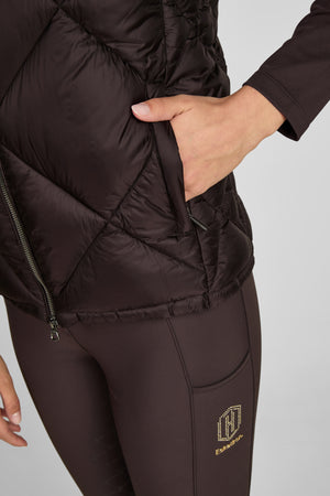 L'image montre une personne portant une veste matelassée noire et des leggings assortis. La veste est en tissu matelassé et semble être rembourrée, offrant une apparence chaude et confortable. Le design comporte des coutures horizontales qui donnent un aspect structuré et élégant. La veste possède une fermeture éclair visible au centre, qui s'étend de haut en bas. Il y a également une poche extérieure avec une fermeture éclair sur le côté droit, dans laquelle la personne a inséré sa main....