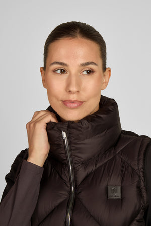 Dans l'image, on peut voir une femme qui pose pour un portrait. Elle est vêtue d'une veste noire matelassée avec une fermeture éclair et un col haut, qui semble être de type coupe-vent. La veste possède un petit logo rectangulaire sur la poitrine, côté gauche, de couleur sombre avec un motif ou texte blanc. En dessous de la veste, elle porte un haut à manches longues de couleur gris foncé. La femme a les cheveux tirés en arrière, coiffés de manière lisse et soignée. Elle a une expression...