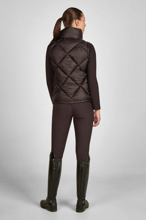 Dans l'image, une femme est photographiée de dos, se tenant dans une posture droite et élégante. Elle porte une tenue qui semble être conçue pour des activités équestres ou de plein air. Elle est vêtue d'un gilet matelassé de couleur sombre, probablement marron ou brun foncé, avec un motif en losanges. Le gilet est ajusté à la taille et semble offrir une bonne isolation thermique. Sous le gilet, elle porte un haut à manches longues, également sombre, qui s'harmonise bien avec le reste de sa...