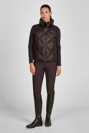 La photo montre une femme debout, vêtue d'une tenue élégante et fonctionnelle qui semble être conçue pour des activités en plein air ou équestres. La femme porte un gilet matelassé avec une coupe ajustée et une fermeture éclair frontale. Le gilet présente un motif en losanges avec des coutures fines qui lui donnent une apparence structurée et sophistiquée. Il est de couleur sombre, probablement marron foncé ou noir. En dessous du gilet, elle porte un haut à manches longues noir qui épouse...