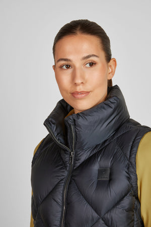 L'image représente une femme vue de face, qui semble être en train de poser pour une photographie. Elle porte un gilet noir matelassé avec une fermeture éclair sur le devant. Le gilet a un design en losanges, ce qui donne une apparence texturée et élégante. Le col du gilet est haut et légèrement plissé, ajoutant une touche de sophistication. Sous le gilet, elle porte un haut jaune moutarde qui contraste avec le gilet noir. Le haut jaune a des manches longues et semble être en tissu lisse et...