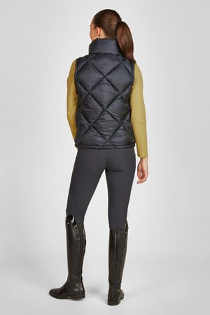 L'image montre une personne vue de dos, vêtue d'une tenue décontractée et élégante. Voici une description détaillée : 1. **Vêtements :** - **Gilet :** La personne porte un gilet noir matelassé avec un motif en losanges. Le gilet est sans manches et semble être bien ajusté, offrant une silhouette élégante. Il est probablement conçu pour offrir chaleur et confort. - **Haut :** Sous le gilet, elle porte un haut à manches longues dans une teinte moutarde, qui contraste bien avec le noir du...