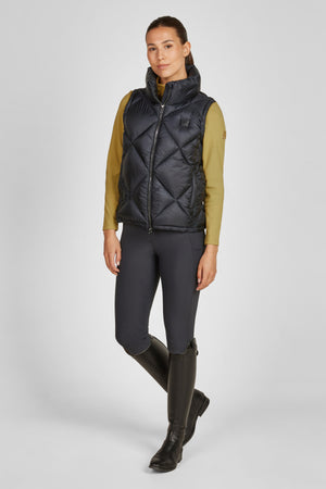 Sur cette image, on peut voir une femme debout, portant une tenue adaptée aux activités de plein air ou à l'équitation. Voici une description détaillée des éléments de sa tenue : 1. **Gilet** : Elle porte un gilet noir matelassé. Ce gilet est doté d'une fermeture éclair centrale. Le matelassage forme des motifs en losanges, conférant un aspect à la fois moderne et sportif. Il possède un col haut, offrant une protection supplémentaire contre le froid. 2. **Chemise** : Sous le gilet, elle...