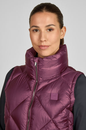 L'image montre une femme regardant directement vers la caméra. Elle porte un gilet matelassé de couleur bordeaux avec une fermeture éclair au centre. Le gilet présente un col haut et un patch ou une étiquette discrète sur le côté gauche de la poitrine, portant un logo ou un nom de marque. En dessous du gilet, elle a une veste ou un pull de couleur foncée, probablement noire ou bleu marine. Ses cheveux sont tirés en arrière, ce qui dégage son visage et met en avant ses traits. Le fond est...
