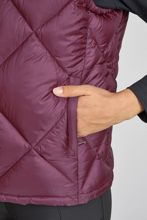 L'image montre une partie d'un vêtement, plus précisément une veste matelassée de couleur pourpre foncé. La veste semble être faite d'un matériau synthétique léger et étanche, probablement destiné à l'isolation thermique. Elle présente un motif de coutures en losange, typique des vestes matelassées, qui forme des poches remplies d'une matière isolante. Sur le côté de la veste, on peut voir une main qui tient une poche latérale. Cette poche est fermée par une fermeture éclair, ce qui permet...