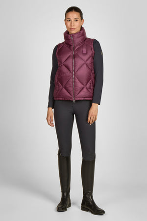 Cette image présente une femme vêtue d'une tenue décontractée mais élégante, avec une touche sportive. Voici une description détaillée : 1. **Gilet matelassé** : La femme porte un gilet matelassé de couleur bordeaux. Ce vêtement est conçu avec des motifs en losanges, typiques des gilets matelassés. Il semble léger et pourrait être en matériau synthétique, probablement pour offrir une isolation thermique tout en restant respirant. Le gilet possède une fermeture éclair frontale qui monte...