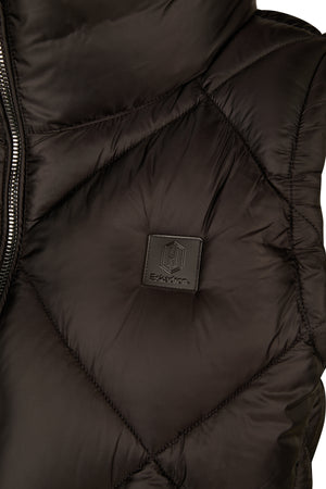 L'image montre une vue rapprochée d'un gilet matelassé noir. Le tissu semble être léger et rembourré, probablement conçu pour offrir une bonne isolation thermique et un confort. Le gilet présente une fermeture éclair visible sur le côté gauche de l'image, avec des dents métalliques et une languette de traction assortie. À l'intérieur du gilet, il y a une étiquette cousue qui est noire avec un logo hexagonal distinct en relief. Le logo contient une initiale stylisée "H" au centre. Sous le...