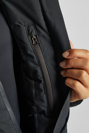 L'image montre un gros plan sur une partie intérieure d'une veste noire. On peut observer une fermeture éclair verticale avec un curseur métallique de couleur argentée. La fermeture éclair semble être située dans une poche intérieure de la veste. Le tissu autour de la fermeture éclair est noir et semble être en matériau synthétique, probablement imperméable ou résistant à l'eau, ce qui est courant pour les vestes de type vêtements d'extérieur. On peut également voir une main humaine sur le...