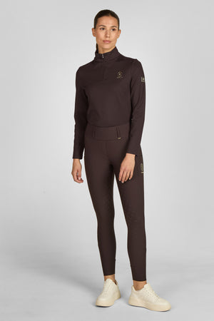 Sur l'image, une femme se tient debout, immobile, contre un fond gris uniforme. Elle est vêtue d'une tenue sportive de couleur marron foncé. Elle porte une veste à manches longues avec une fermeture éclair partielle au niveau du col. La veste est ajustée et possède des logos ou inscriptions sur le côté gauche de la poitrine et sur la manche droite. Le bas de sa tenue est composé de leggings moulants, également marron foncé, qui vont jusqu'à ses chevilles. Ces leggings présentent des motifs...