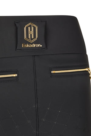 L’image montre une partie d’un pantalon d’équitation de la marque Eskadron. Voici une description détaillée : 1. **Marque et Logo** : - Au niveau de la taille, un emblème distinctif est cousu sur le tissu. - Cet emblème est constitué d’un motif hexagonal avec les lettres "H" et "E" en son centre. - Le nom "Eskadron" est inscrit en dessous de l’emblème en lettres dorées. 2. **Couleur et Matériau** : - Le pantalon est principalement noir. - Le matériau semble être un tissu...