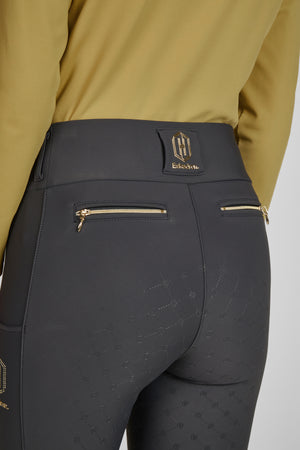 L'image montre une vue rapprochée de la partie inférieure du corps d'une personne portant des jodhpurs noirs. Ces jodhpurs sont dotés de deux poches zippées sur l'arrière, chacune avec une fermeture éclair dorée. La matière des jodhpurs semble être extensible et confortable, et elle est ornée d'un motif en relief répété. Ce motif est constitué de petits carrés formant des losanges et comportant le logo de la marque "Eskadron". Au niveau de la taille, il y a une bande de tissu noir avec une...