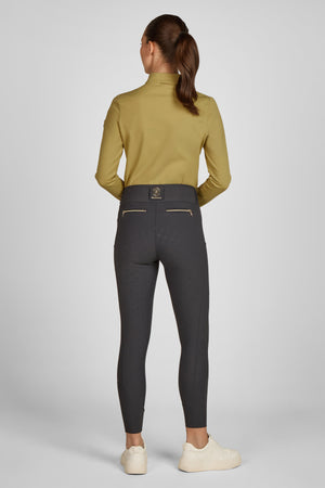 L'image représente une femme vue de dos, se tenant debout dans une posture droite. Elle porte une tenue qui semble être adaptée à une activité sportive ou équestre. Voici une description détaillée de son apparence et de ses vêtements : 1. **Cheveux** : Ses cheveux sont attachés en une queue de cheval basse et soignée. 2. **Haut** : Elle porte un haut long à manches longues de couleur moutarde, qui semble être ajusté et légèrement extensible, probablement pour offrir une liberté de...