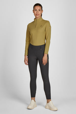 L'image montre une femme debout, vêtue d'une tenue sportive. Elle porte un haut à manches longues de couleur olive avec une fermeture éclair partielle au col. Le haut est ajusté et présente un logo sur la poitrine ainsi que sur les manches. Elle est également vêtue d'un pantalon d'équitation noir et moulant, avec des coutures au niveau des genoux et de la taille, accentuant la forme du vêtement. Le pantalon comporte une ceinture noire avec un logo visible sur la boucle. Elle porte des...