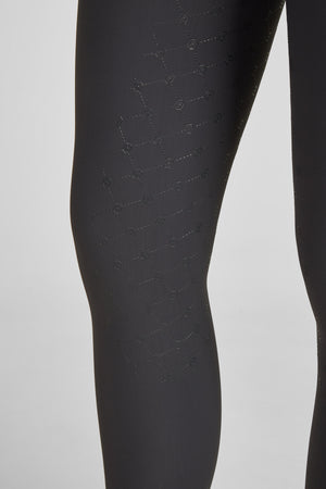 L'image présente une vue rapprochée des jambes d'une personne vêtue d'un pantalon ou de leggings noirs. Le tissu du vêtement est orné d'un motif texturé distinctif composé de petits losanges imbriqués les uns dans les autres. Chaque losange contient un petit motif de "G" stylisé, répété de manière uniforme sur toute la surface visible du tissu. Le fond de l'image est blanc, ce qui contraste fortement avec le noir du pantalon, mettant ainsi en valeur le motif texturé. Les jambes sont...