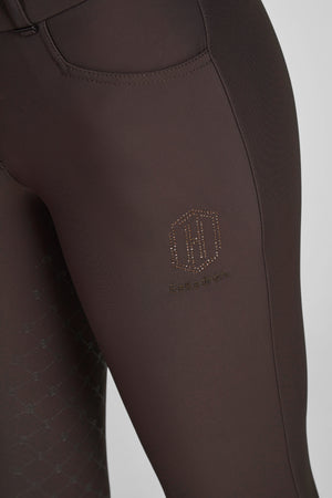 L'image montre une partie d'une jambe de pantalon d'équitation, probablement conçue pour le sport équestre. Le pantalon est de couleur marron foncé et présente un design élégant et fonctionnel. À l'arrière de la cuisse, on peut voir un logo distinctif composé de deux lettres "H" stylisées, formées par des petits points ou perforations, probablement pour ajouter une touche de ventilation et de style. En dessous de ce logo, le mot "Eskadron" est inscrit en lettres cursives, indiquant la...
