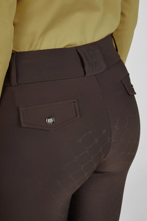 L'image montre la partie inférieure d'une personne vêtue de pantalons d'équitation bruns. Ces pantalons sont conçus avec des détails spécialement adaptés à l'équitation, tels que le tissu extensible pour assurer confort et flexibilité. Voici les éléments visibles : 1. **Ceinture et Taille :** - Une ceinture en tissu est attachée autour de la taille. - La ceinture présente un logo avec les lettres "HH" et le mot "Eskadron" écrit en vert. - La taille des pantalons est bien ajustée...