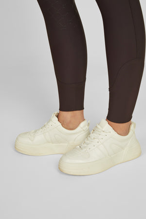 L'image montre les jambes d'une personne portant des vêtements de sport et des chaussures de style décontracté. Les jambes sont couvertes de collants noirs moulants avec des détails brillants et des sections texturées ou cousues. Les collants sont conçus pour offrir un soutien et une flexibilité, probablement utilisés pour des activités sportives telles que l'équitation ou le fitness. Les chaussures sont de couleur crème et présentent un design lisse avec des motifs perforés sur le côté....