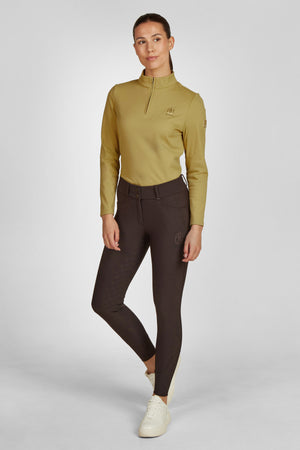 Dans l'image, une femme se tient debout contre un fond blanc uni. Elle est vêtue d'une tenue décontractée mais élégante. Elle porte un haut à manches longues de couleur moutarde, qui est ajusté et comporte une fermeture éclair au col. Ce haut semble confortable et adapté à une activité physique légère. En bas, elle porte un pantalon d'équitation marron foncé qui épouse parfaitement ses formes, mettant en valeur sa silhouette. Le pantalon est orné de deux poches sur l'avant et d'un petit logo...