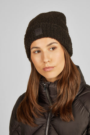 Sur cette image, on voit une personne portant un bonnet noir en tissu moelleux et un manteau noir. Le bonnet est épais et douillet, adapté pour les périodes froides. Il présente un logo ou une étiquette carrée sur le devant, légèrement sur le côté. La personne a des cheveux longs et bruns qui tombent librement sur ses épaules. Son expression est neutre et elle regarde directement l'objectif de la caméra. Le manteau qu'elle porte semble être une veste matelassée, conçue pour offrir une...