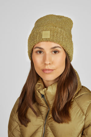 L'image représente une femme portant un bonnet d'hiver en tissu bouclé de couleur beige ou vert olive clair. Le bonnet est bien ajusté et couvre ses oreilles, offrant une protection contre le froid. Il comporte un petit emblème carré cousu sur le devant, probablement une marque ou un logo. La femme a de longs cheveux lisses et bruns qui tombent sur ses épaules. Elle porte également une veste beige ou vert olive clair, apparemment matelassée et légère, avec une fermeture éclair visible sur le...