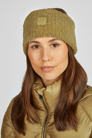 L'image représente une femme regardant directement l'objectif de la caméra. Elle porte un serre-tête en tissu bouclé dans une teinte vert olive. Le serre-tête est orné d'une petite étiquette carrée en cuir avec un logo ou un emblème discret, probablement représentant la marque "ELK" ou quelque chose de similaire. La femme a des cheveux longs et bruns qui tombent sur ses épaules. Elle porte également une veste matelassée en nylon vert olive avec une fermeture éclair visible. L'arrière-plan est...