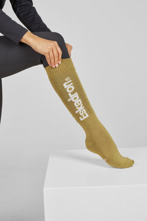 Dans cette image, on peut voir une personne assise sur un banc blanc, avec une jambe tendue vers l'avant. La personne porte une chaussette montante de couleur kaki. La chaussette est ornée du mot "Eskadron" écrit en lettres blanches verticalement le long de la jambe. La personne porte également un pantalon noir et a posé ses mains sur son genou plié. Le fond est simple et blanc, mettant en valeur la chaussette et le pantalon. La posture est décontractée et naturelle, suggérant un contexte...