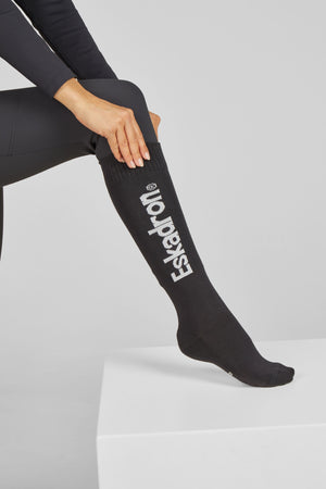 L'image montre une personne assise sur une surface blanche, vêtue de leggings noirs. La personne porte des chaussettes noires qui montent jusqu'aux genoux. Les chaussettes sont ornées de l'inscription "Eskadron" en lettres blanches verticales, avec un petit logo au-dessus du texte. La main gauche de la personne repose sur son genou, légèrement fléchi, accentuant les chaussettes. L'arrière-plan est uniformément blanc, ce qui met en valeur les vêtements sombres et les chaussettes. La...