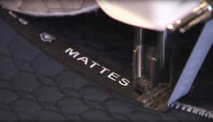 Machine à coudre fixant un galon marqué MATTES sur le bord d'un tapis matelassé bleu nuit.